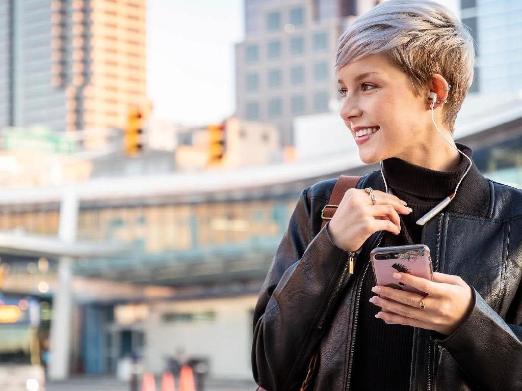 زن جوان در حال استفاده از هندزفری بلوتوث گردنی و بررسی اتصال آن به گوشی